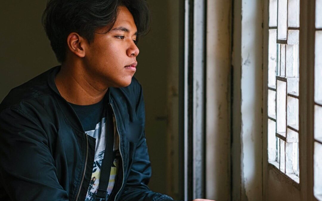 A young man gazes out of a window, deep in thought, reflecting on his experiences. A Fort Worth trauma therapist can help process past trauma through trauma therapy in Fort Worth.