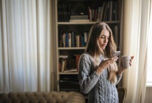 A woman in a cozy sweater looks at her phone with a tense expression, holding a coffee cup in her other hand. Relationship trauma can create lasting emotional distress, making it difficult to feel safe and trust again. A Fort Worth trauma therapist can provide support, and trauma counseling in Fort Worth, TX can help process past experiences and build healthier connections.