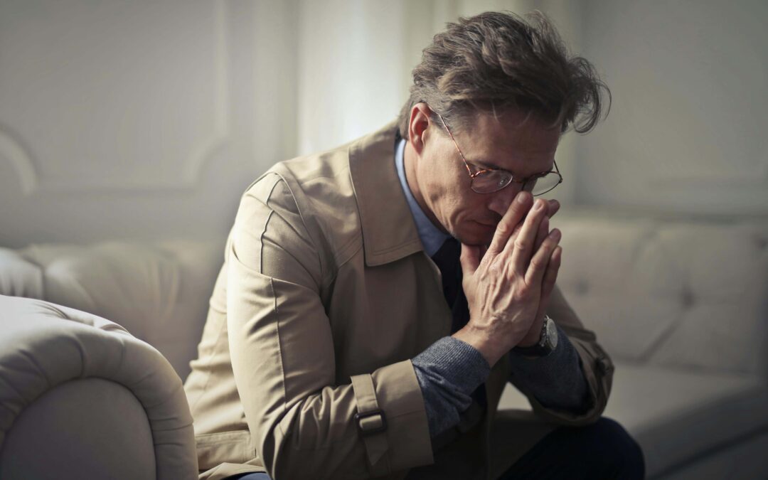 A middle-aged man sits on a couch with his head bowed and hands clasped, appearing deep in thought. He looks distressed, reflecting the emotional toll of past relationship trauma. Seeking trauma therapy in Fort Worth, TX or working with a therapist for relationship trauma in Fort Worth, TX can help process these lingering effects and support healing.