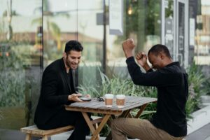 Two men sit at an outdoor café table, one celebrating joyfully while the other smiles and looks at his phone. Healing from relationship trauma is possible with the right support. Trauma therapy in Fort Worth, TX can help process past wounds, and a therapist for relationship trauma in Fort Worth, TX can guide you toward rebuilding trust and connection.